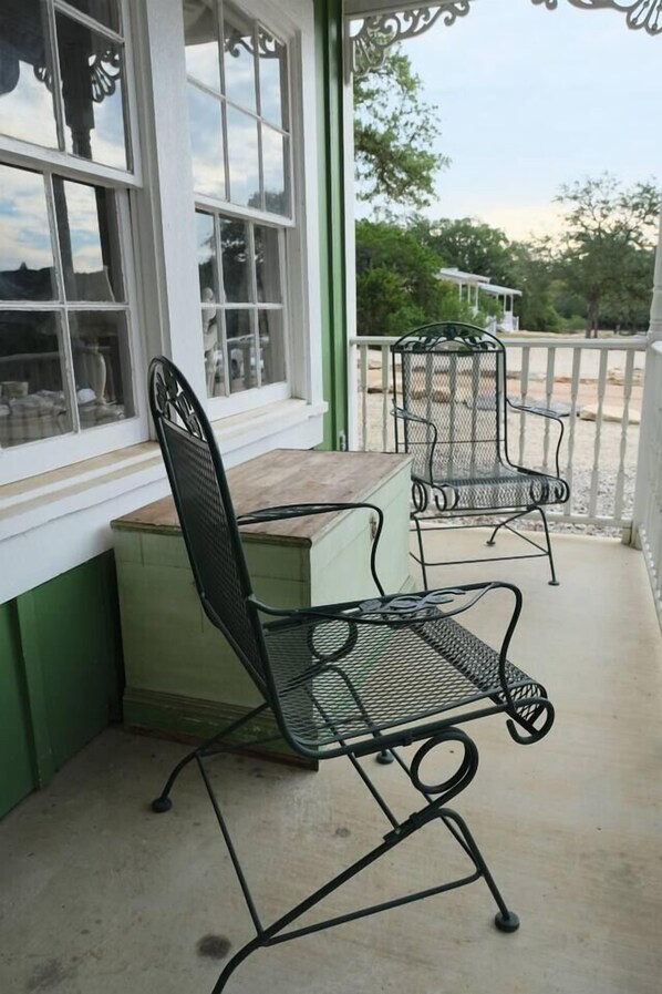 Uma área de estar coberta ao ar livre, com cadeiras e mesa de metal, uma janela com vista para árvores e um corrimão branco.