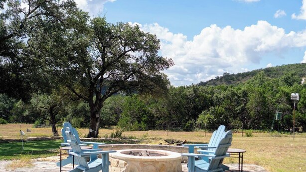 A fire pit area with two Adirondack chairs, surrounded by a stone fire pit, set in a grassy field with trees and a hill in the background.