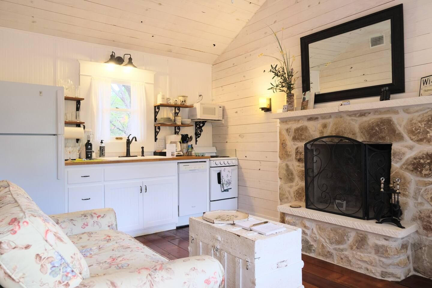 Un salon chaleureux avec un canapé à motifs floraux, une cheminée en pierre, un poêle blanc et une cuisine dotée de meubles blancs et d’un plan de travail en bois.