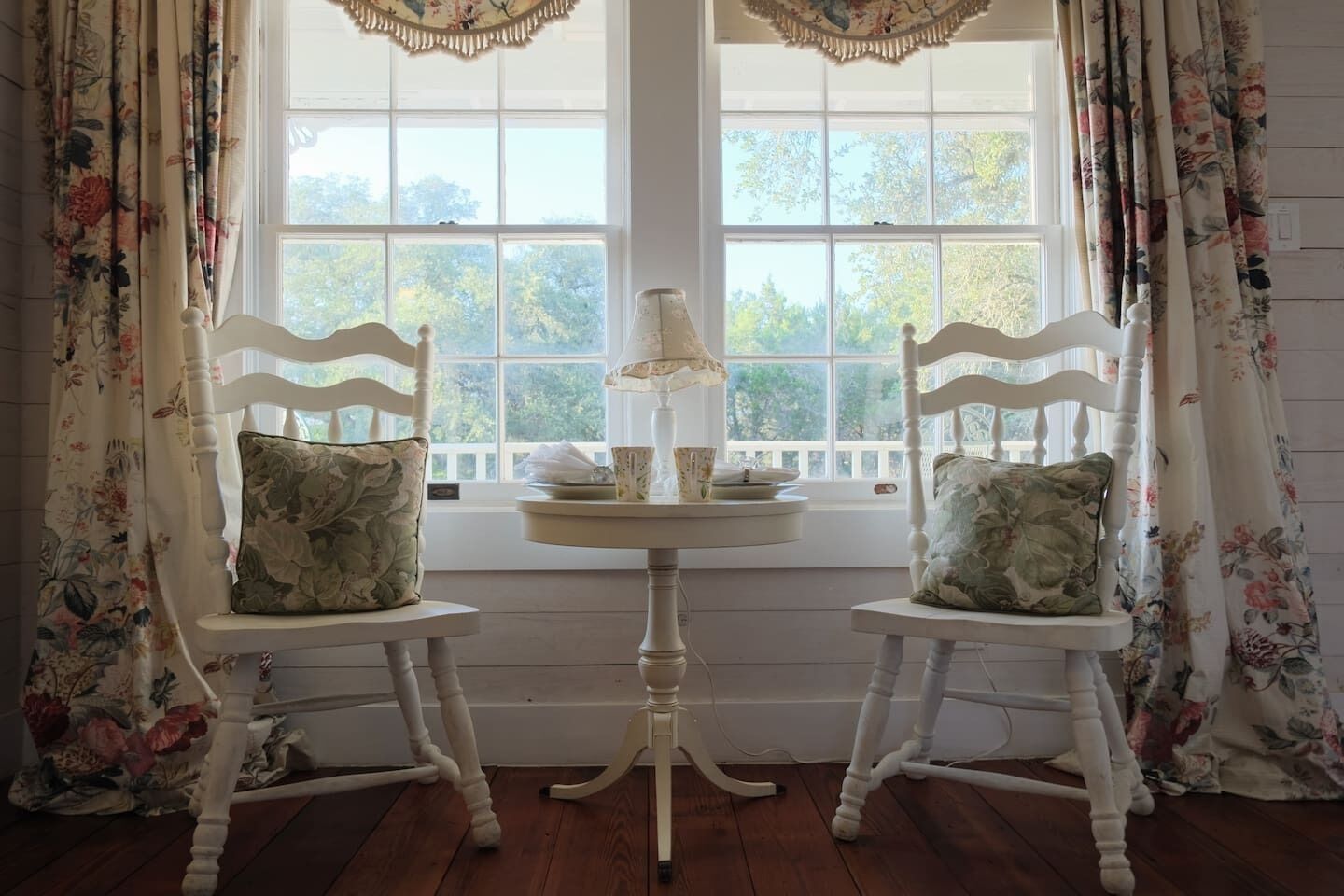 Deux chaises blanches avec des coussins à motifs floraux, une petite table ronde avec une lampe et une fenêtre ornée de rideaux à fleurs.