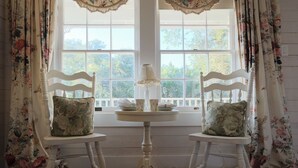Two white chairs with floral cushions, a small round table with a lamp, and a window with floral curtains.