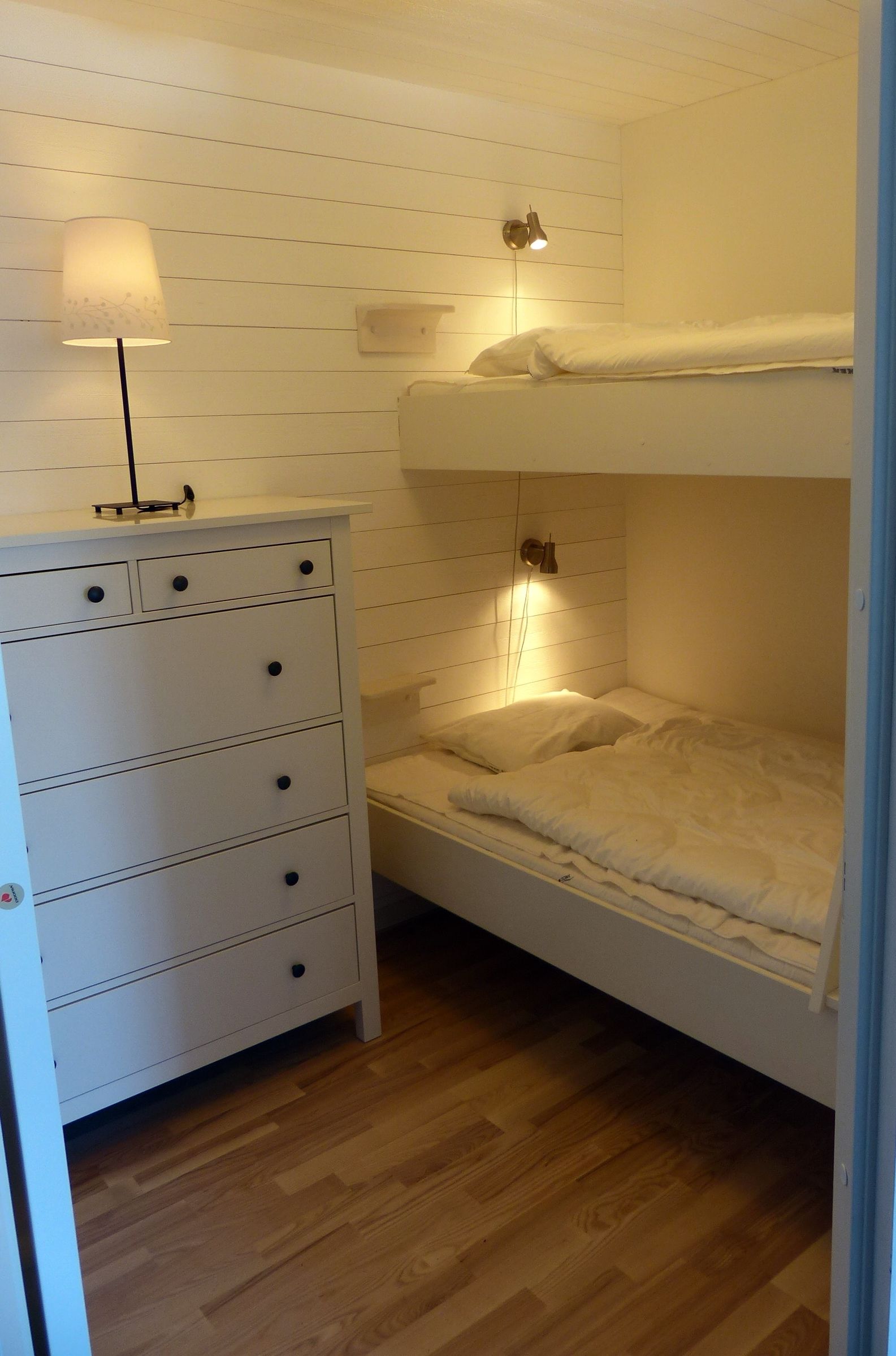 Een stapelbed met nachtkastje en lamp, houten vloer en een witte dressoir met lades.