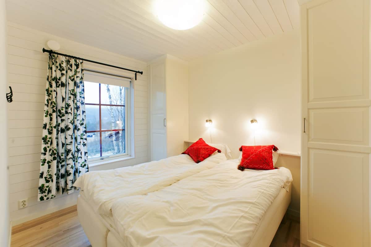 A bedroom with a bed, two red pillows, a window with curtains, and a wall-mounted light fixture.