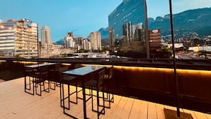 A rooftop terrace with tables and chairs, overlooking a cityscape with modern buildings and mountains in the background. - Auténtico Monterrey (San Pedro Garza García)