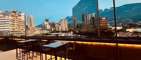 A rooftop terrace with tables and chairs, overlooking a cityscape with modern buildings and mountains in the background.