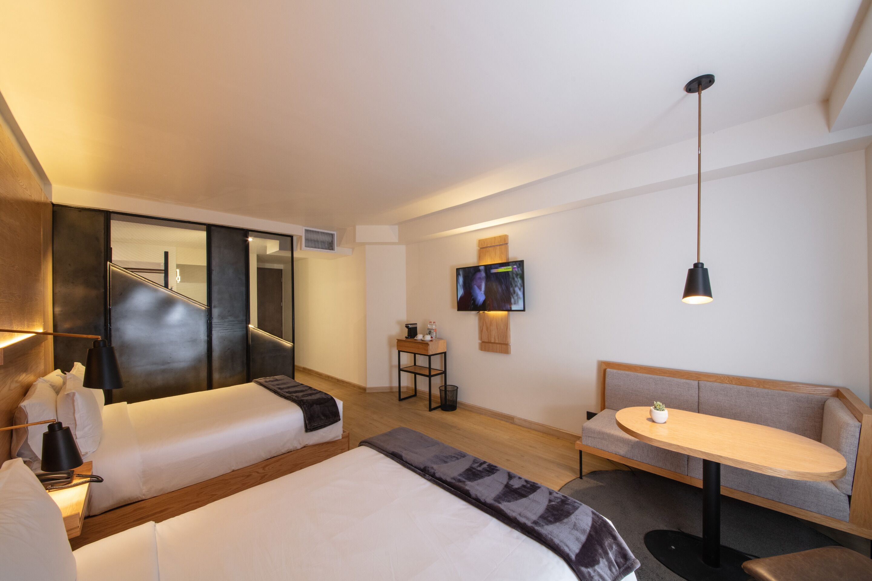 Una habitación de hotel moderna con una cama grande, un sofá, una mesita pequeña, un televisor montado en la pared y una lámpara de techo.