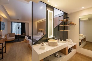 Habitación de hotel moderna con una cama grande, una zona de estar con sofá y mesa, un lavamanos redondo con espejo y un armario con perchero.