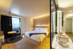 A modern hotel room with a large bed, a bedside table, a chair, a desk, a large window with curtains, and a bathroom with a sink and mirror.