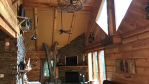 A cozy cabin interior with a stone fireplace, wooden walls, a ceiling fan, and a large window. - Luxury Lake Chalet Hot Tub Private Boat Slip  Amazing Mountain View’s Sleeps 10 (Butler)