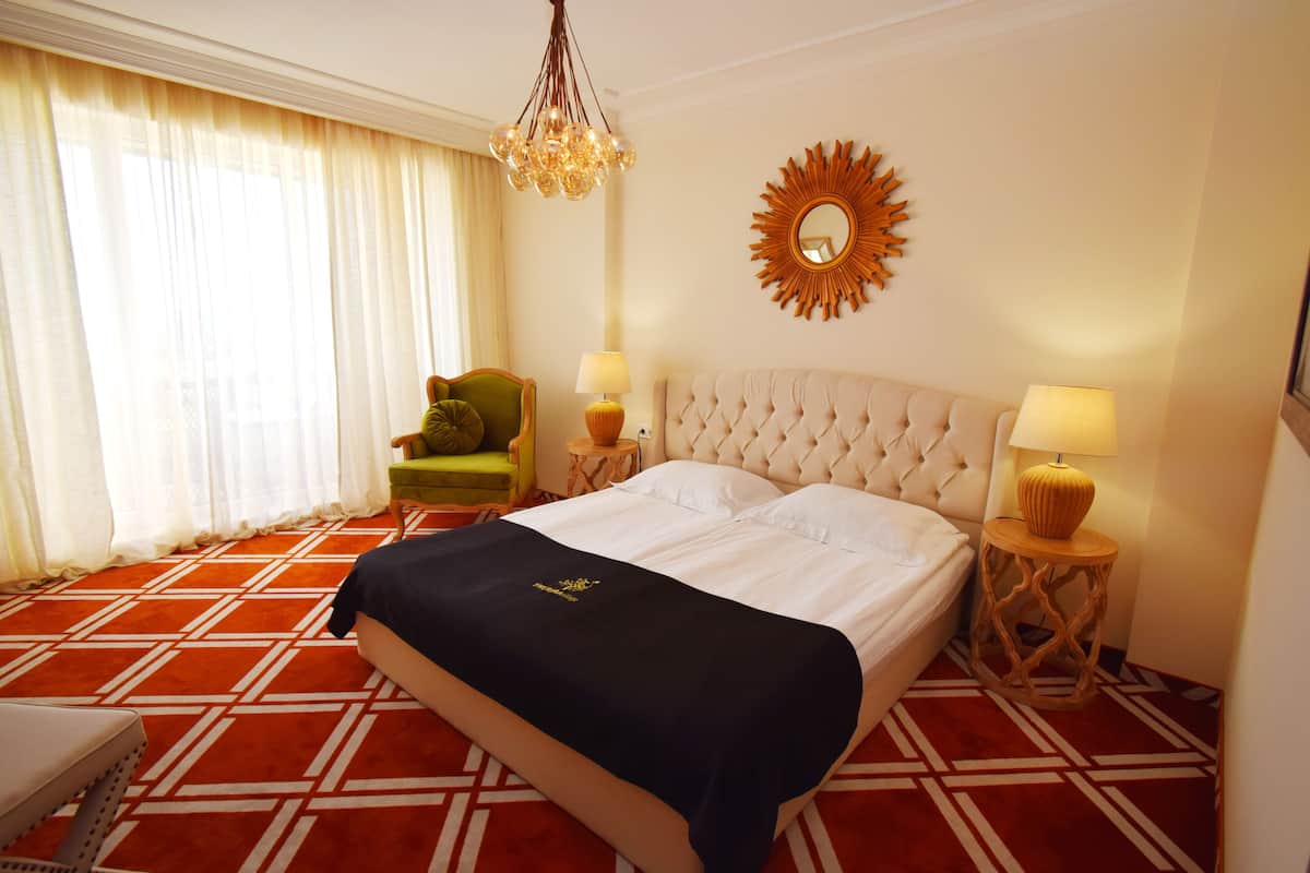 A hotel room with a large bed, two bedside tables with lamps, a green armchair, a large mirror, and a patterned carpet.