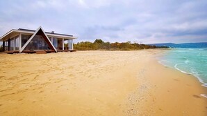 Ett modernt strandhus med en stor glasfasad, beläget på en sandstrand med utsikt över havet och avlägsna kullar.