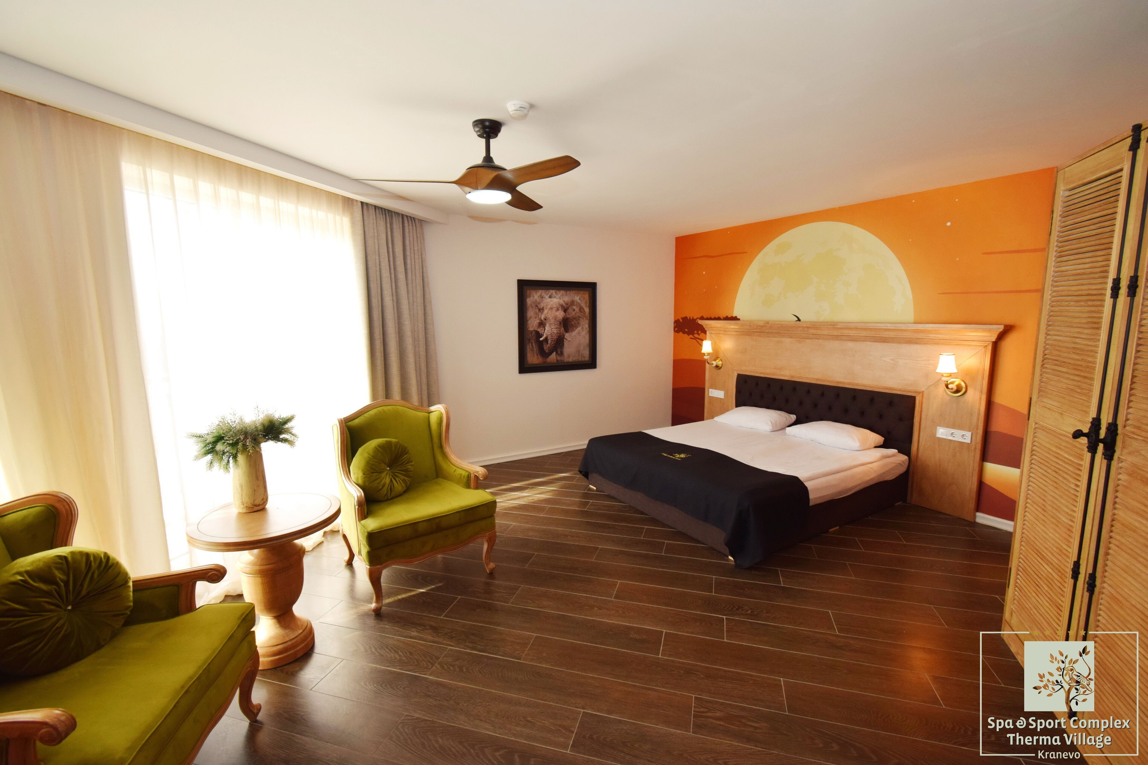 A hotel room with a bed, two green armchairs, a round wooden table with a plant, a ceiling fan, and a wall mural.