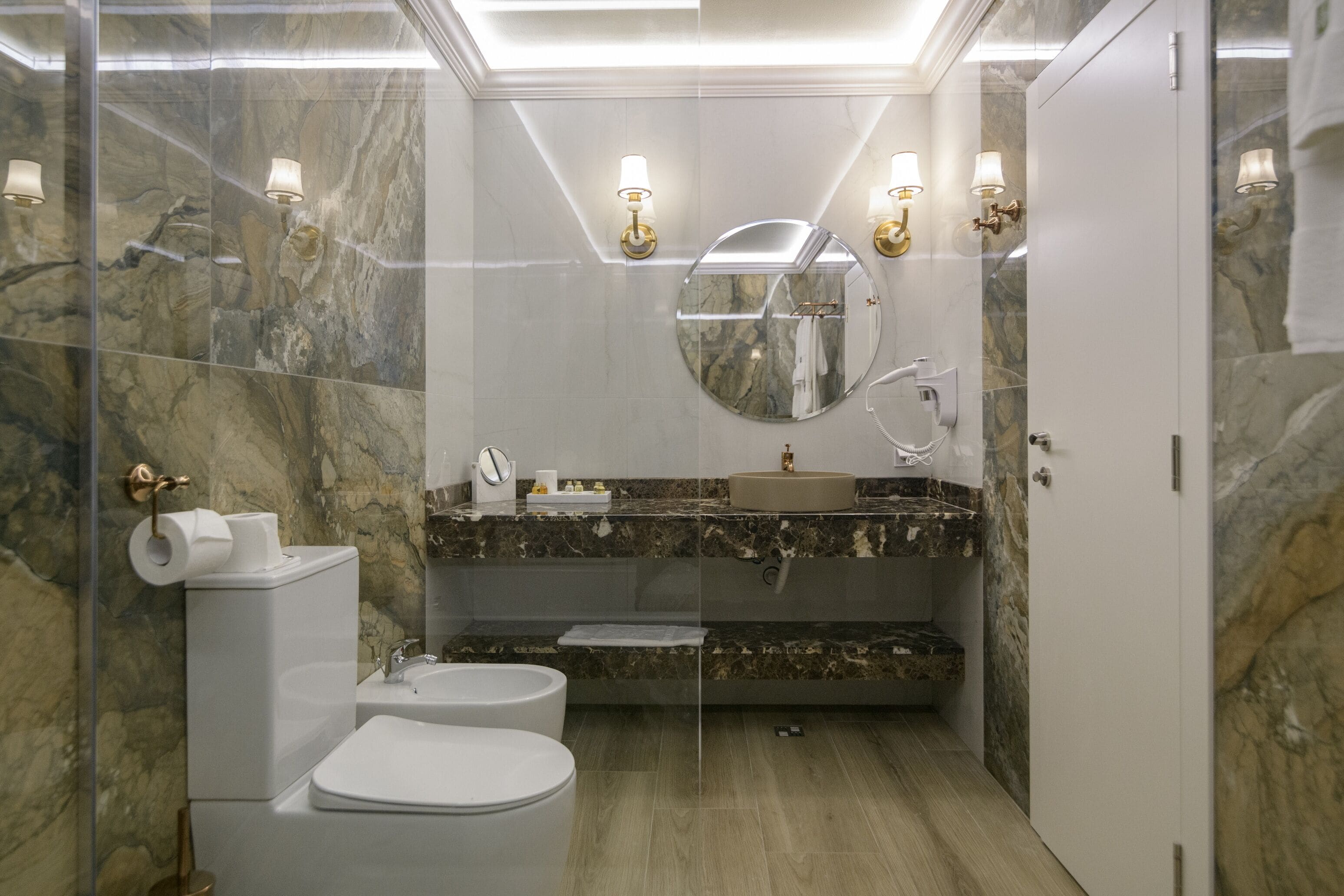 A modern bathroom with a marble wall, a round mirror, a sink, and a toilet.