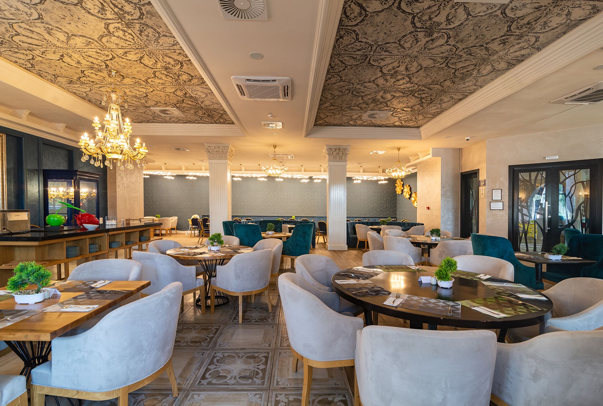 Un restaurant spacieux avec de hauts plafonds, des motifs décoratifs élaborés et d’élégants lustres. Les tables sont garnies de chaises blanches et de petites plantes en pot.
