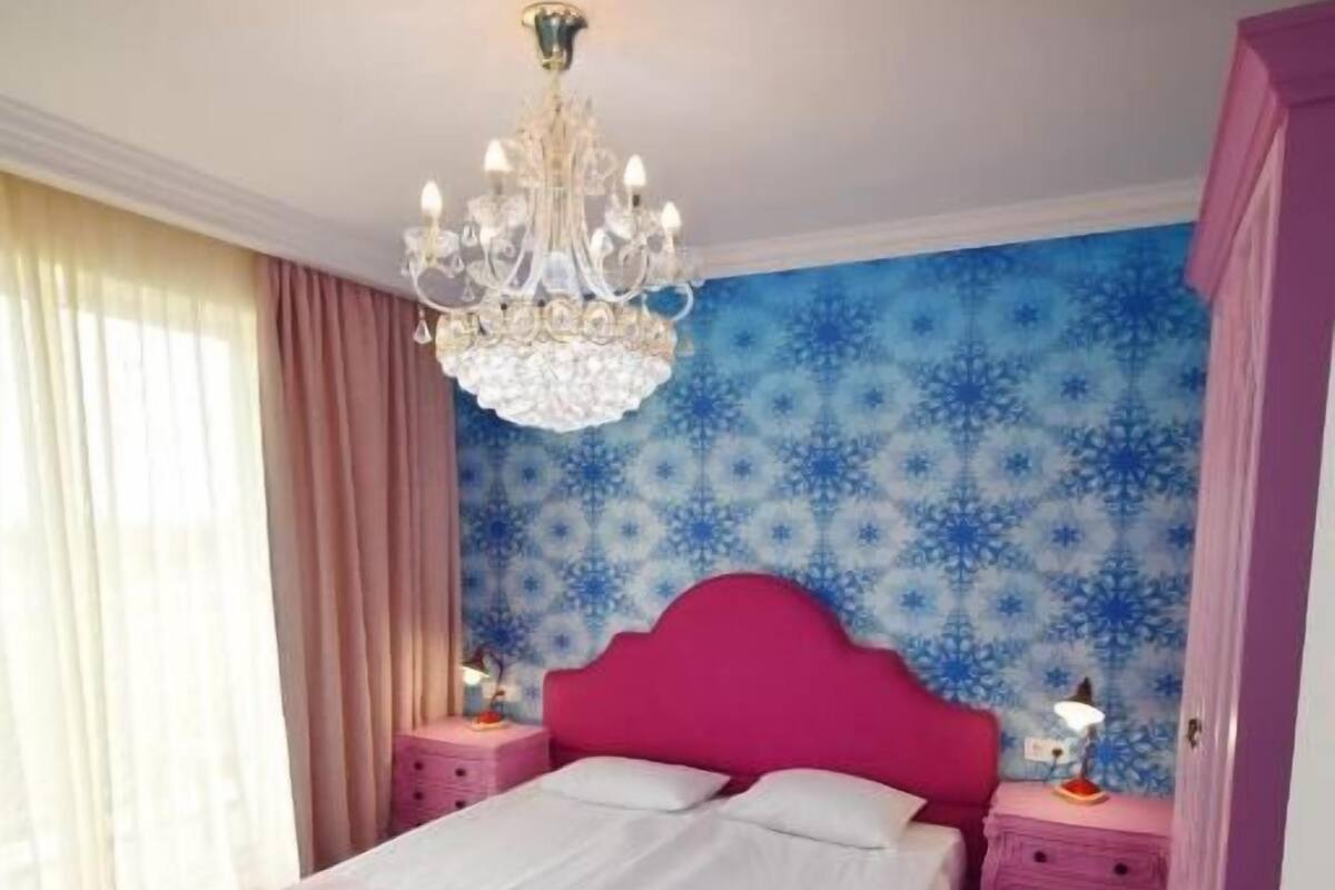 A bedroom with a pink bed, a chandelier, a nightstand with a lamp, and a blue patterned wall.