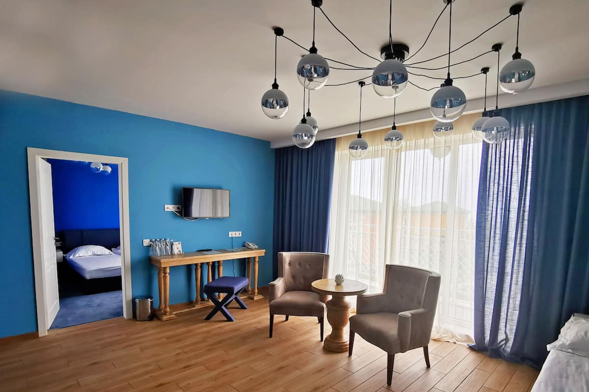 A hotel room with a blue wall, wooden flooring, a desk with a computer, two armchairs, a small round table, and a TV mounted on the wall.