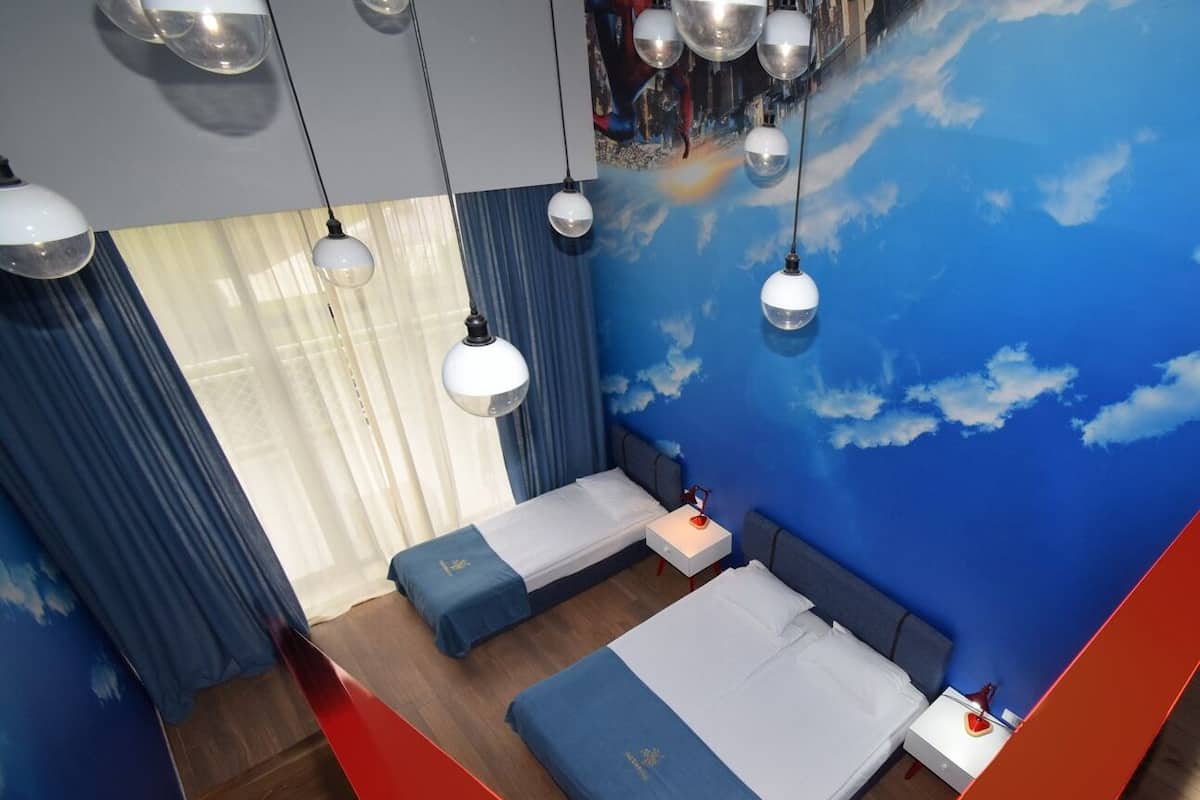 A modern hotel room with two beds, a large window with curtains, and a mural of clouds on the ceiling.