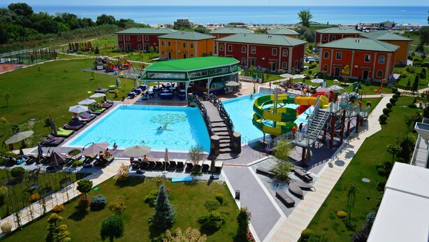 Ett semesterkomplex med en stor swimmingpool, en vattenrutschkana och flera byggnader med gröna tak.