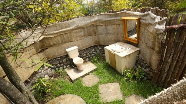 A rustic outdoor bathroom with a toilet and a wooden bench, surrounded by natural stone and greenery.