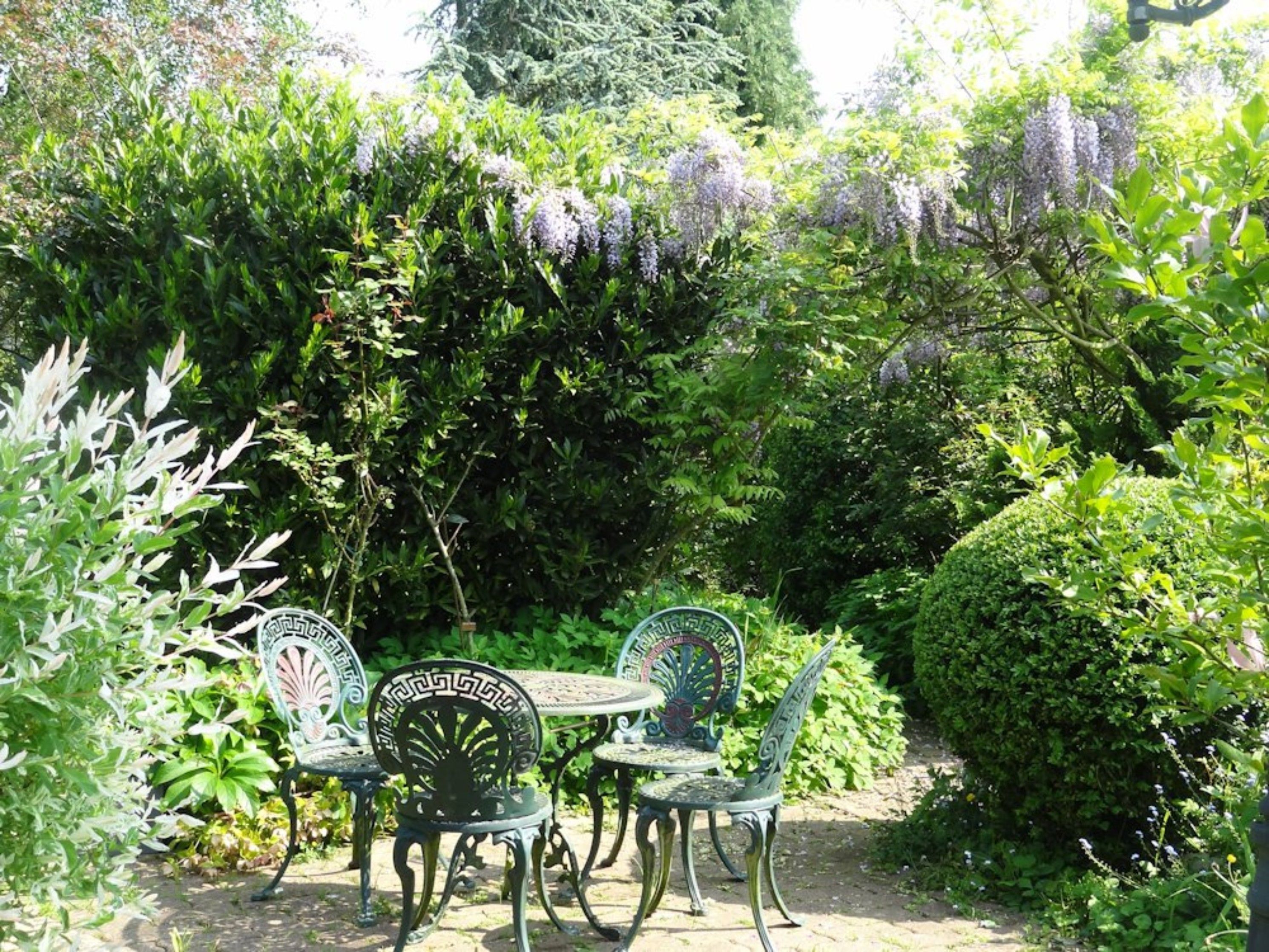 Un jardin extérieur avec une table et quatre chaises, entouré d’une végétation luxuriante et de plantes en fleurs.