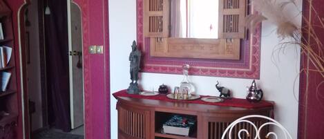 Una habitación con un tocador de madera, un espejo, una cama con colcha roja y dorada, y una silla de metal blanca.