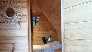 A wooden cabin bedroom with a bed, a small stool, a wall-mounted dreamcatcher, and a window with a view of trees. - Le Frêne Fringant (Sainte-Opportune-la-Mare)