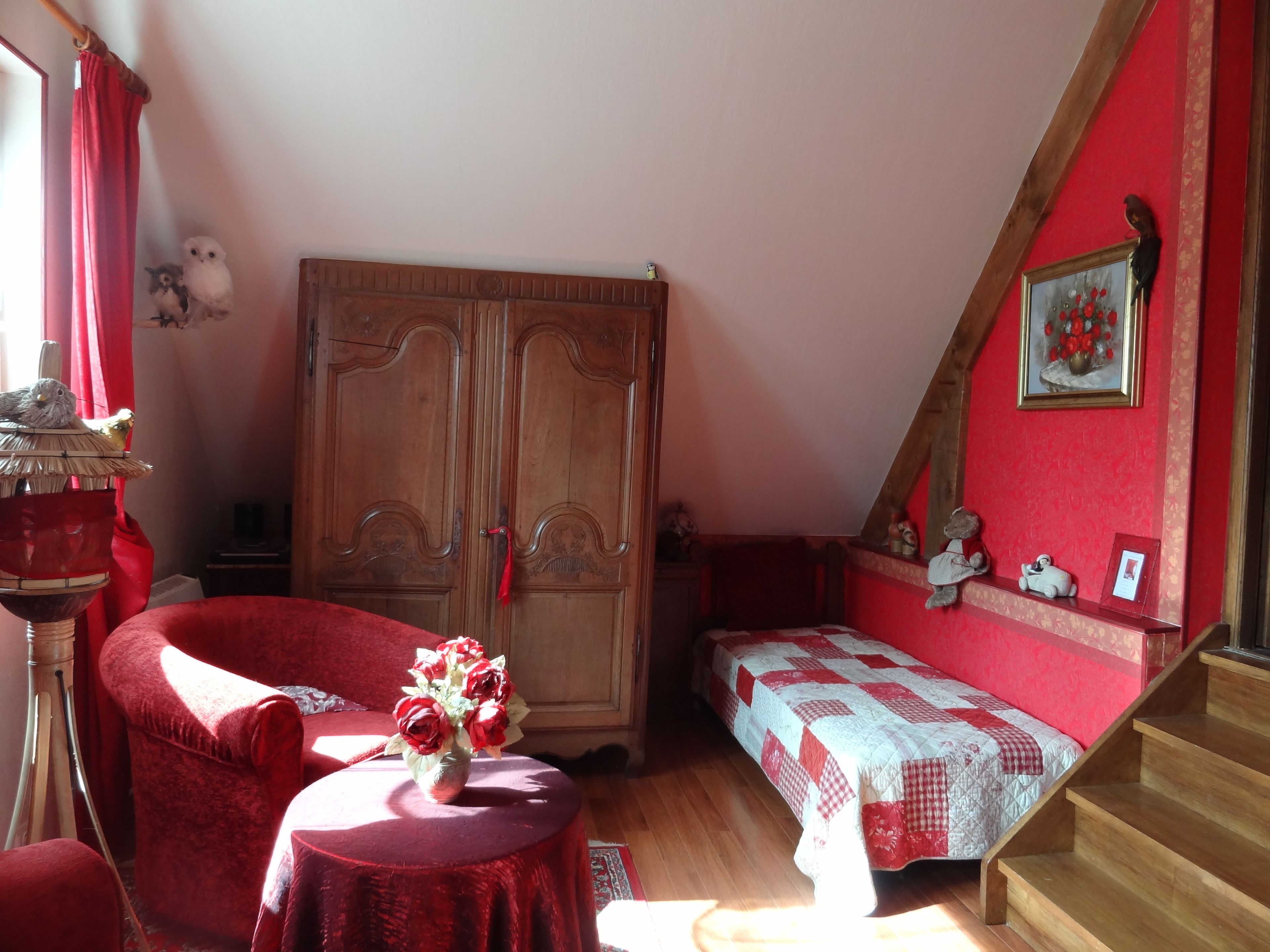 A cozy room with a wooden wardrobe, a red armchair, a small round table with a vase of flowers, a bed with a checkered quilt, and a staircase.