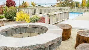 A stone fire pit surrounded by wooden stools in an outdoor setting with a pool and a wooden deck in the background. - Wine Suite at Rellik House. Winery & Alpaca Farm (Central Point)