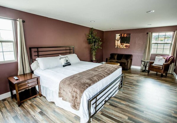 A bedroom with a large bed, a nightstand, a chair, a small table, and a potted plant.