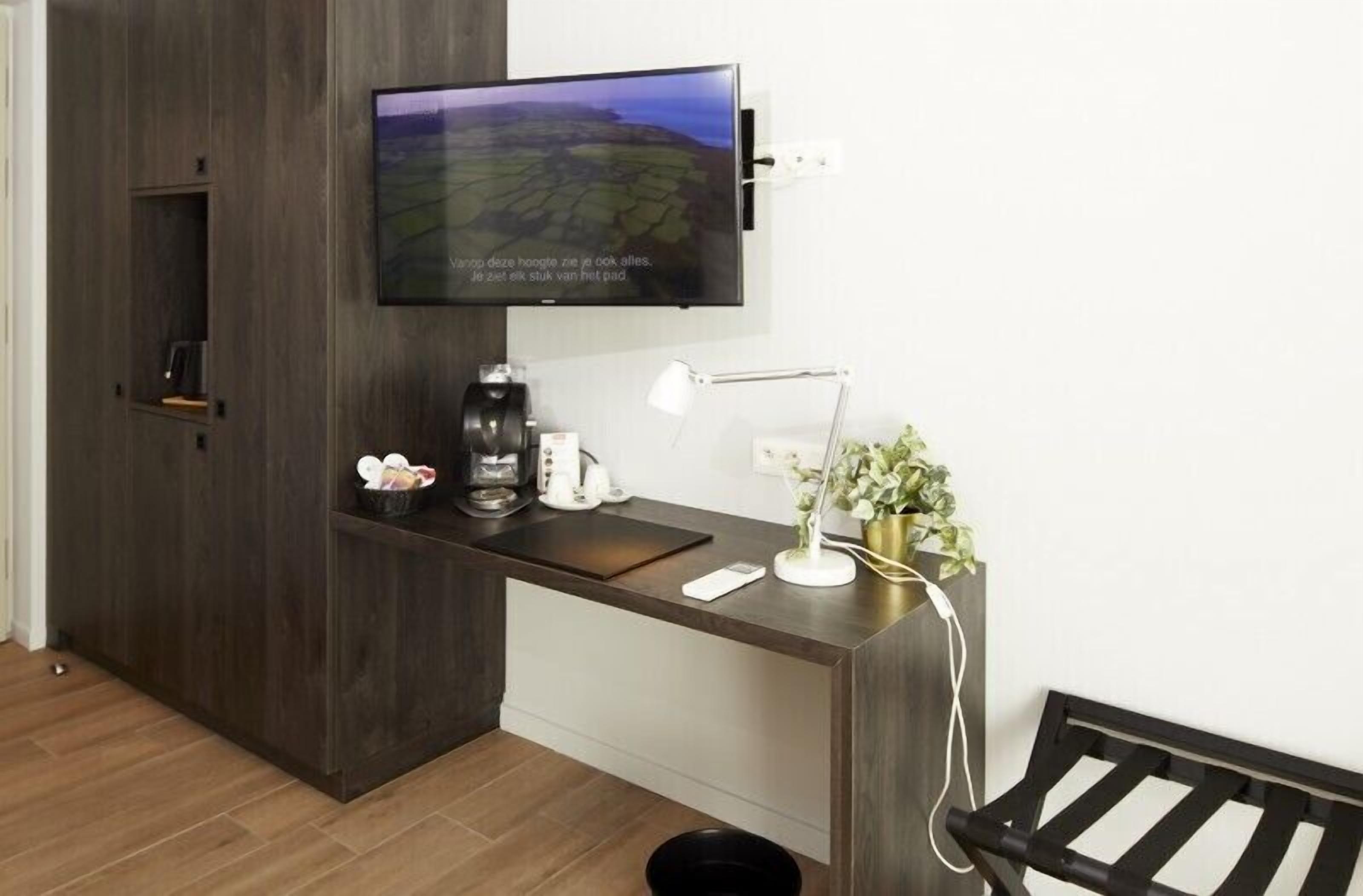 Ett modernt rum med ett skrivbord i mörkt trä, en väggmonterad TV, en kaffebryggare, en lampa och en liten växt.