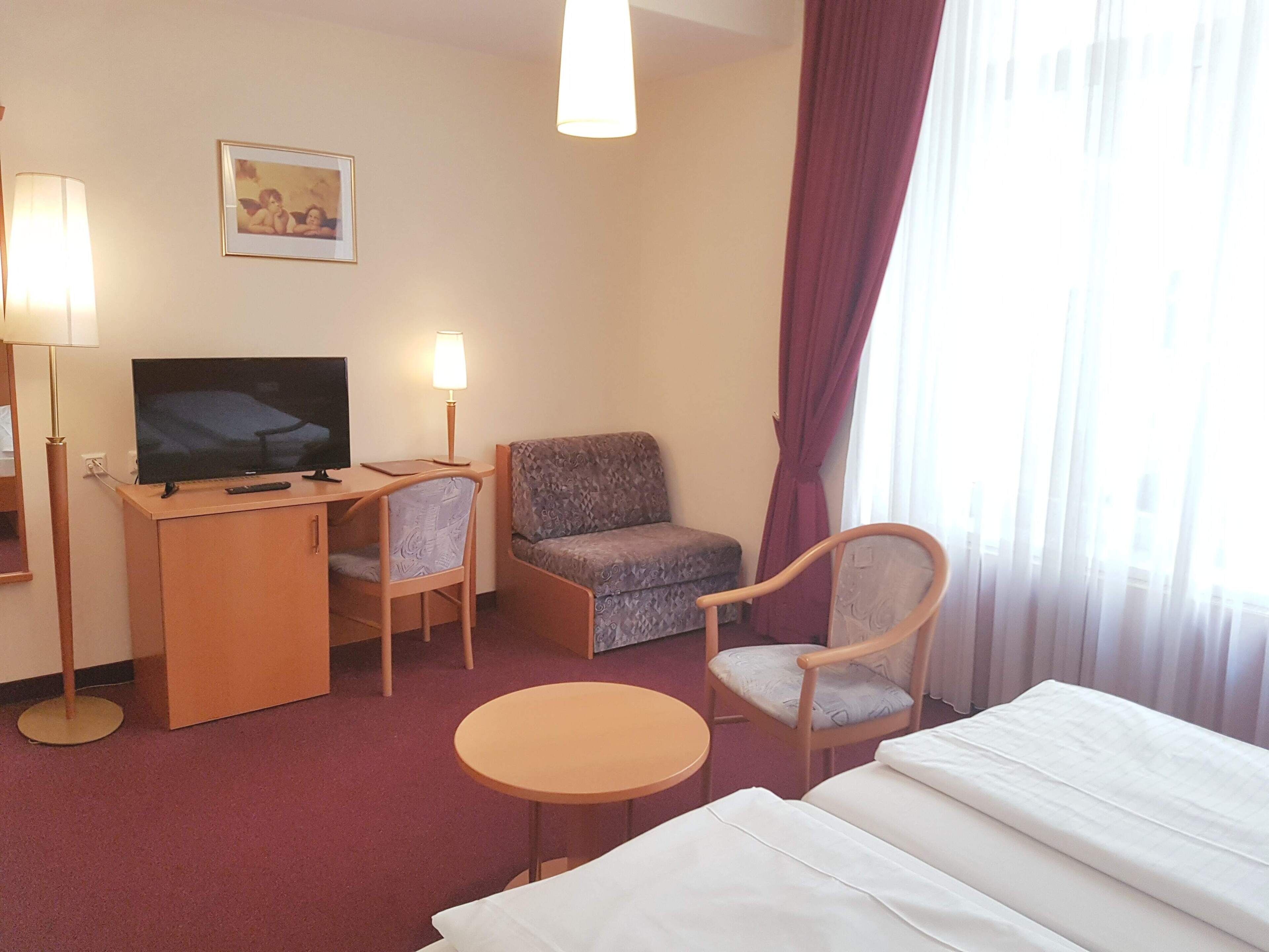 Habitación de hotel con cama, escritorio con televisor, dos sillas, una mesita y una ventana con cortinas.