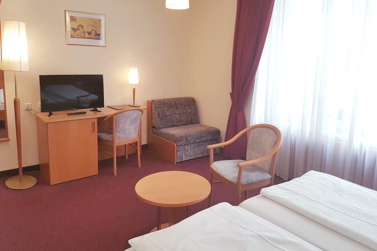 Une chambre d’hôtel comprenant un lit, un bureau avec une télévision, deux chaises, une petite table et une fenêtre avec des rideaux.