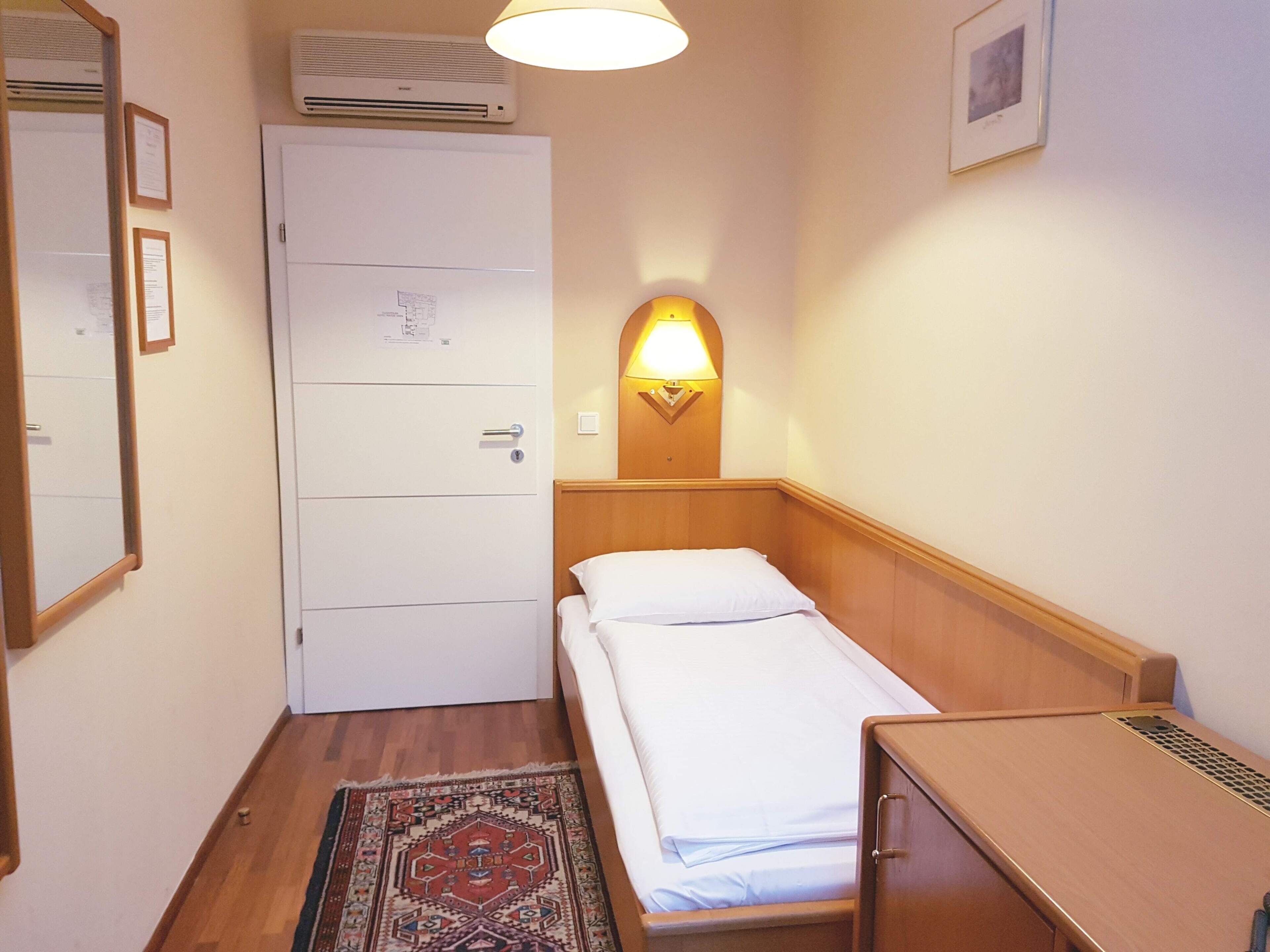 Una habitación de hotel pequeña con una cama individual, una mesita de noche de madera, un escritorio, una lámpara de pared, una puerta con cerradura y una alfombra estampada.