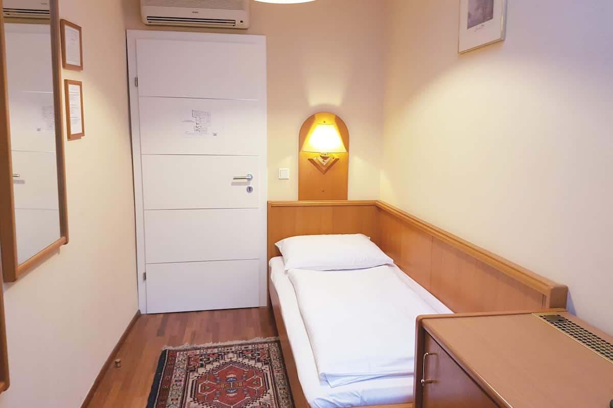 Une petite chambre d’hôtel avec un lit simple, une table de chevet en bois, un bureau, une lampe fixée au mur, une porte avec une serrure et un tapis à motifs.