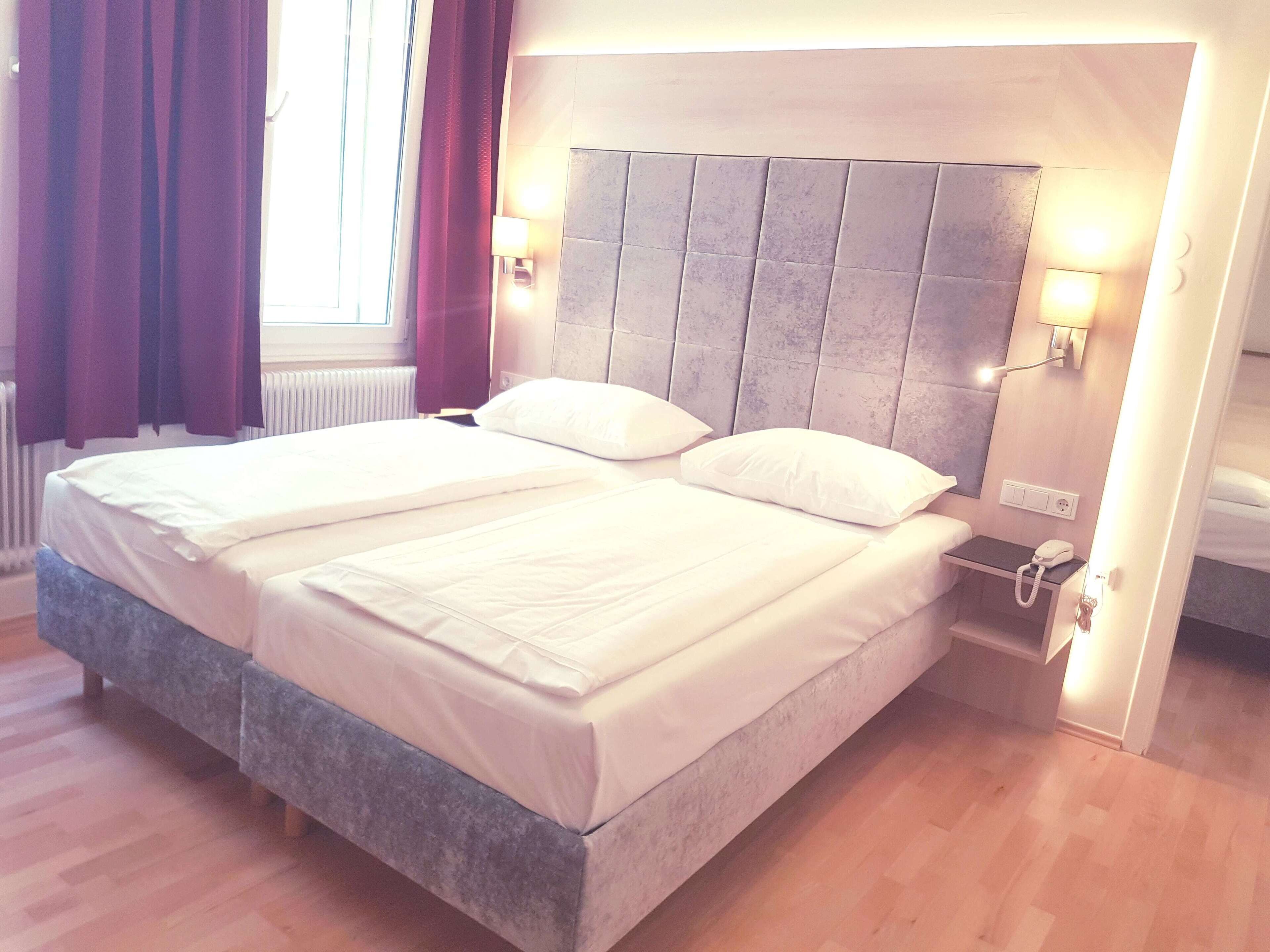 Una cama doble con cabecera gris, ropa de cama blanca y dos lámparas de noche.