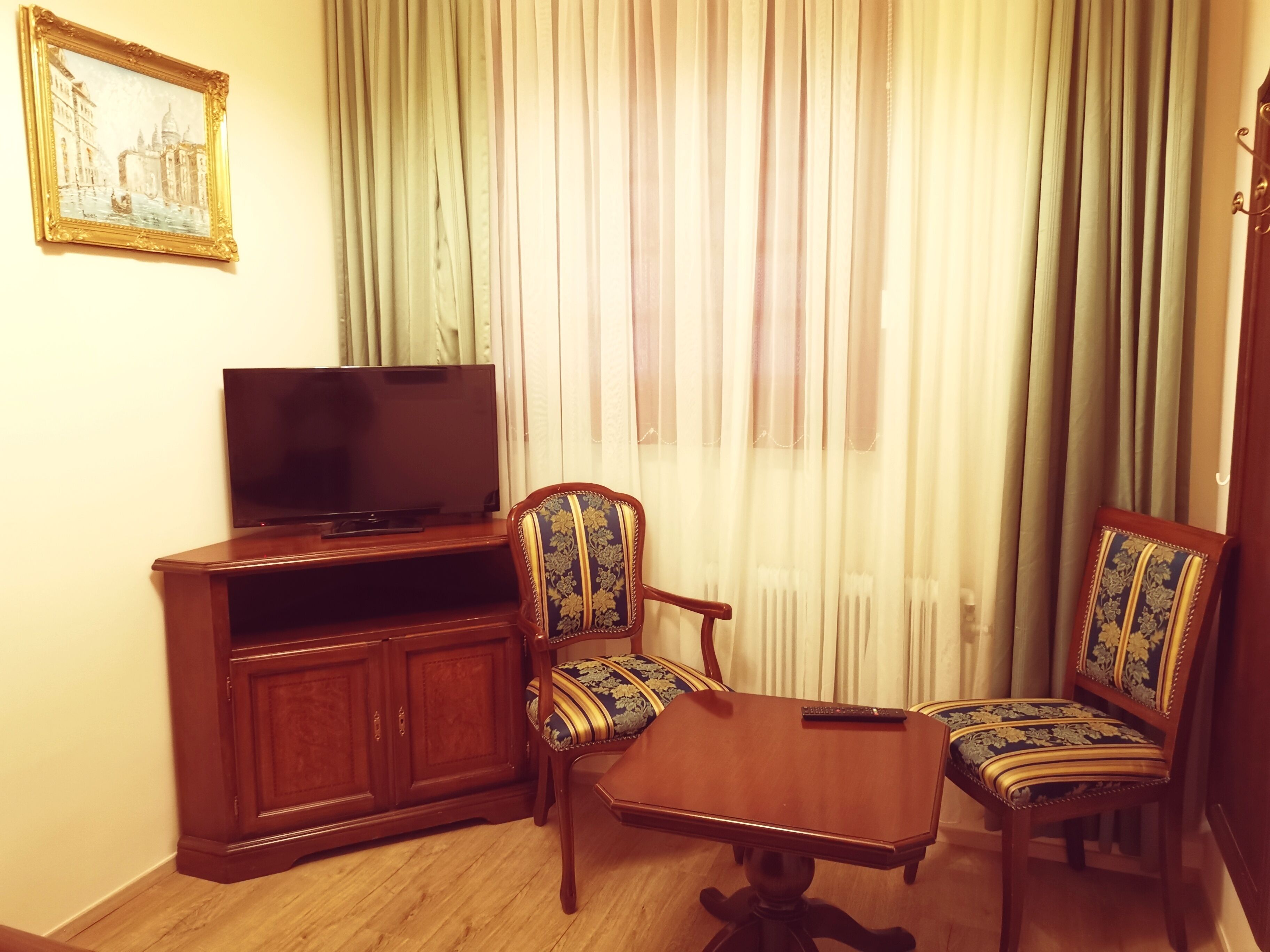 Una habitación con una televisión sobre un soporte de madera, dos sillones tapizados con estampados florales, una mesita de madera y un ventanal con cortinas traslúcidas.