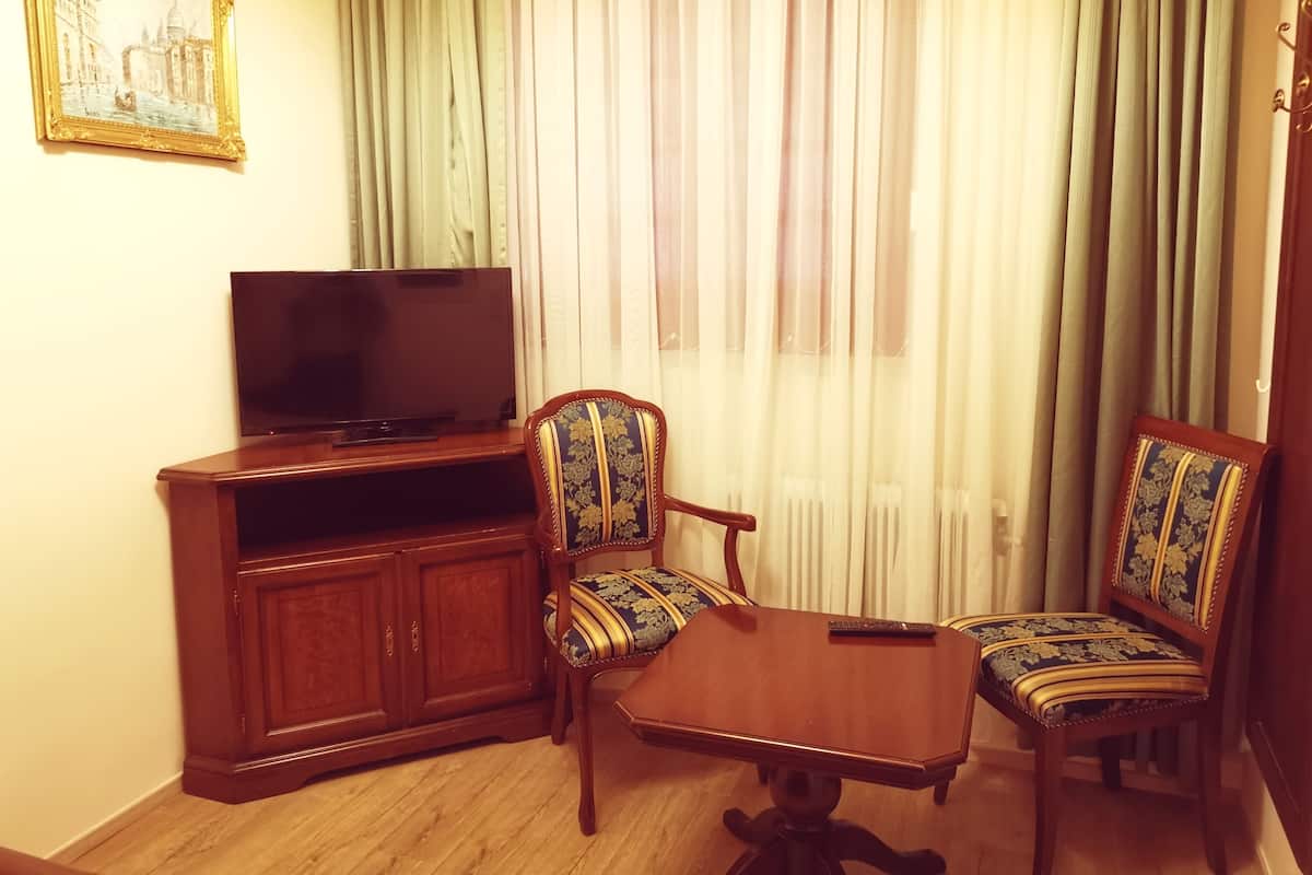Une pièce avec un téléviseur posé sur un meuble en bois, deux fauteuils rembourrés ornés de motifs floraux, une petite table en bois et une grande fenêtre avec des rideaux légers.