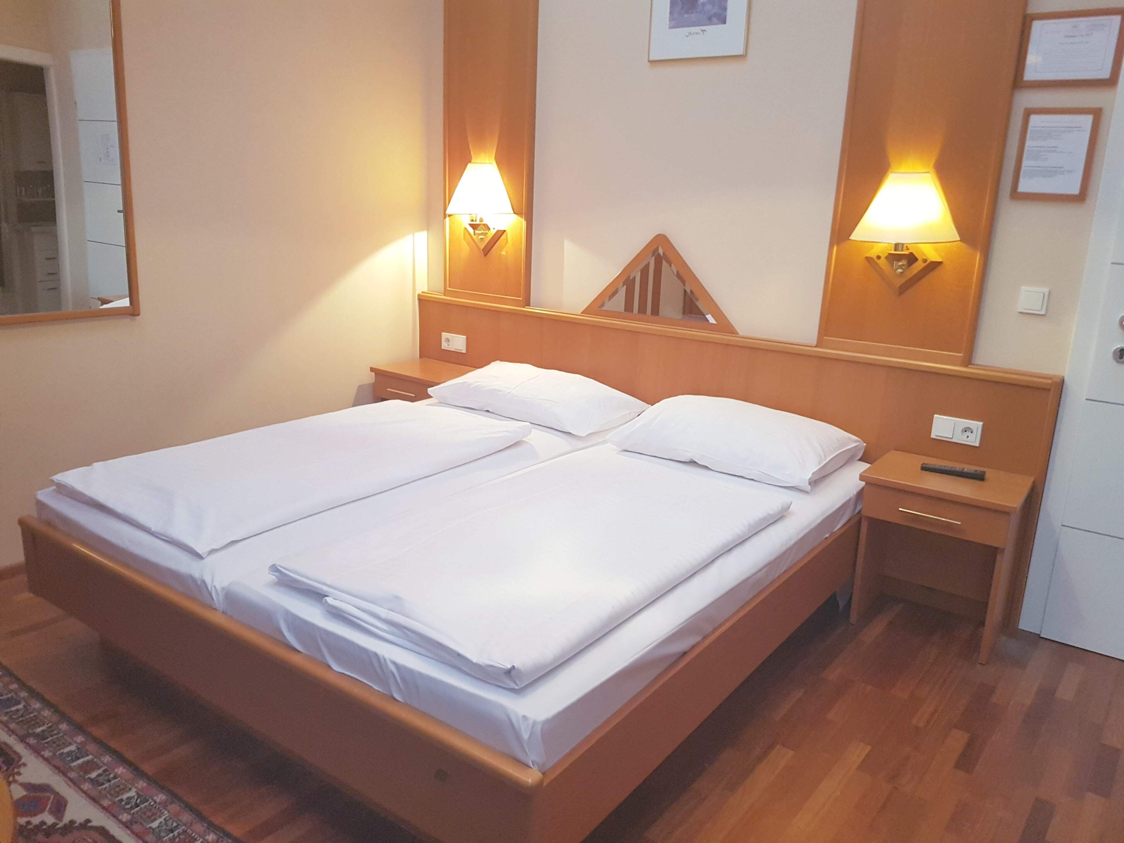 Una cama doble con ropa de cama blanca, dos mesitas de noche, un cabecero de madera, lámparas fijadas a la pared y una mesita auxiliar pequeña con un control remoto.