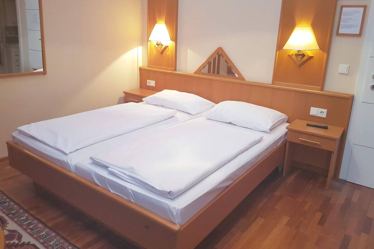 Un lit double avec du linge de lit blanc, deux tables de chevet, une tête de lit en bois, des lampes fixées au mur et une petite table de nuit avec une télécommande.
