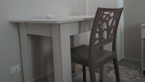 Una habitación pequeña y sencilla con un escritorio y silla blancos, suelo estampado y una lámpara de pared.