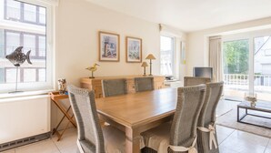 A dining area with a wooden table and wicker chairs, a small cabinet, and two framed pictures on the wall. - Monet (Koksijde)