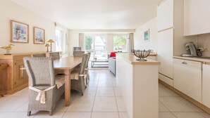 A modern kitchen with white cabinets, a dining area with wicker chairs, and a view through the glass doors. - Monet (Koksijde)