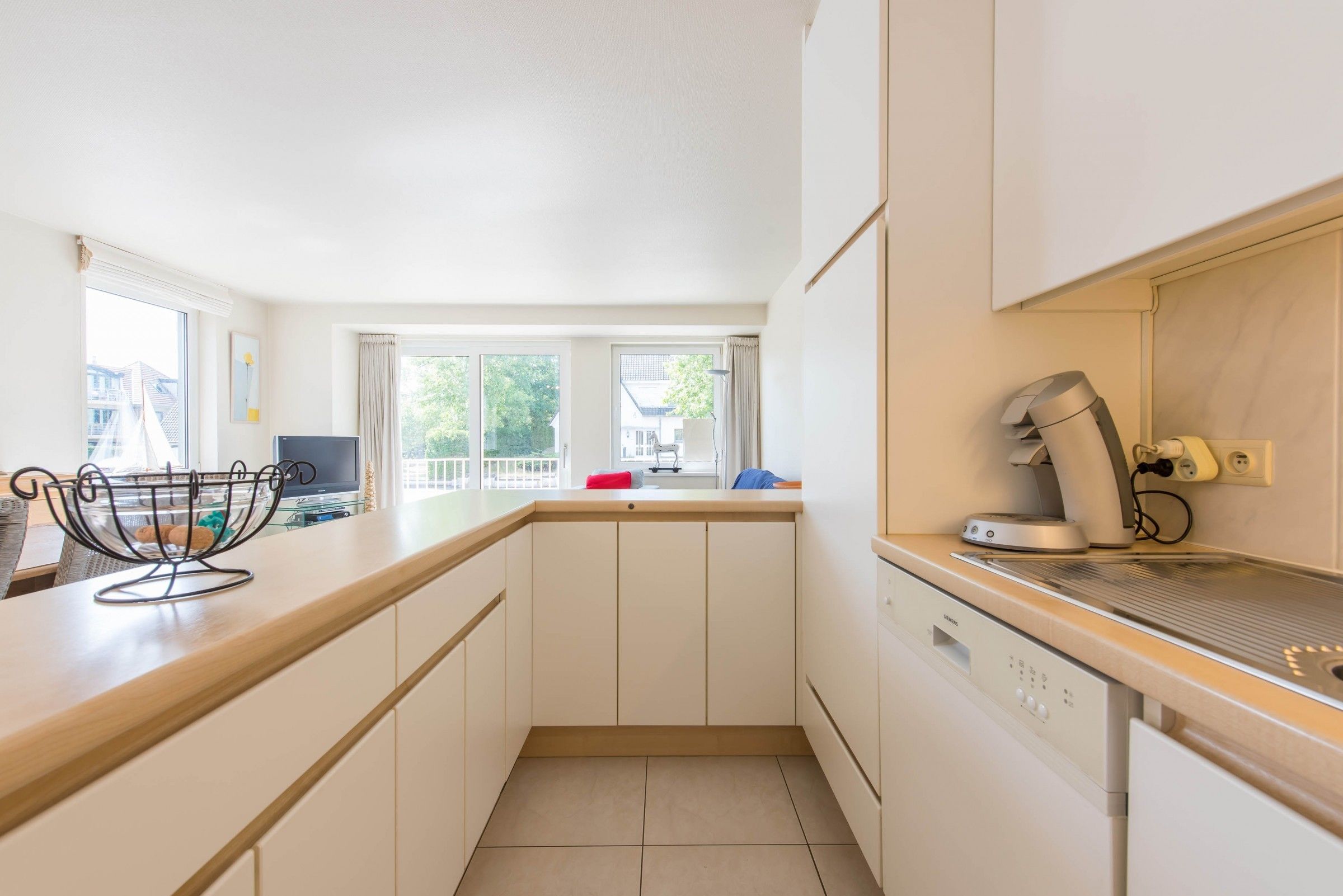A modern kitchen with white cabinets, a built-in dishwasher, and a window overlooking a balcony.