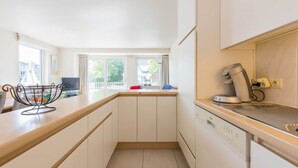 A modern kitchen with white cabinets, a built-in dishwasher, and a window overlooking a balcony. - Monet (Koksijde)