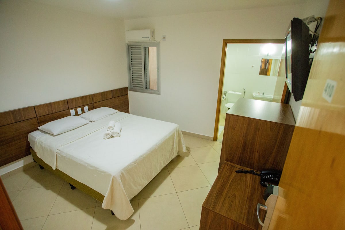 Habitación de hotel con cama, cabecero de madera, ventana con contraventanas, baño visible a través de una puerta abierta y teléfono sobre una encimera de madera.