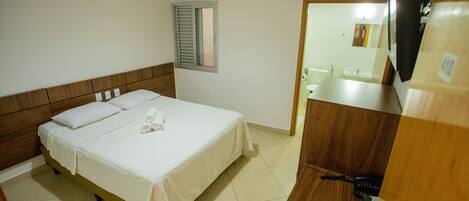 A hotel room with a bed, a wooden headboard, a window with shutters, a bathroom visible through an open door, and a telephone on a wooden counter.