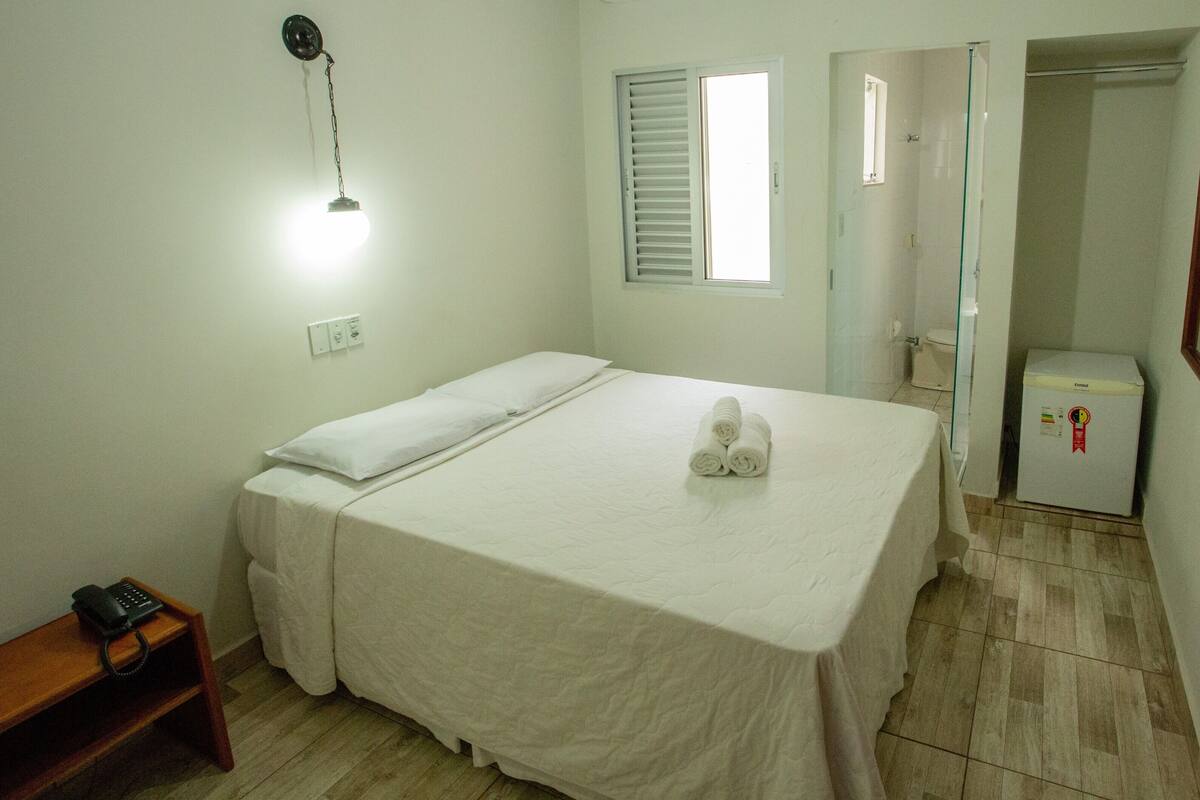 Habitación de hotel con cama, mesita de noche con teléfono, lámpara de pared, ventana con persianas, zona de ducha y refrigerador.
