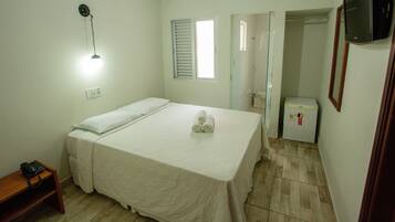 Quarto de hotel com cama, mesa de cabeceira com telefone, luminária na parede, janela com persianas, área de chuveiro e um frigorífico.