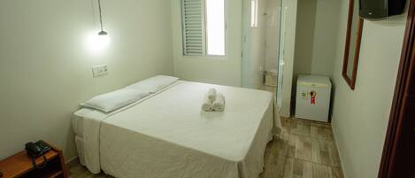 Een hotelkamer met een bed, een nachtkastje met telefoon, een wandlamp, een raam met zonwering, een douchegedeelte en een koelkast.
