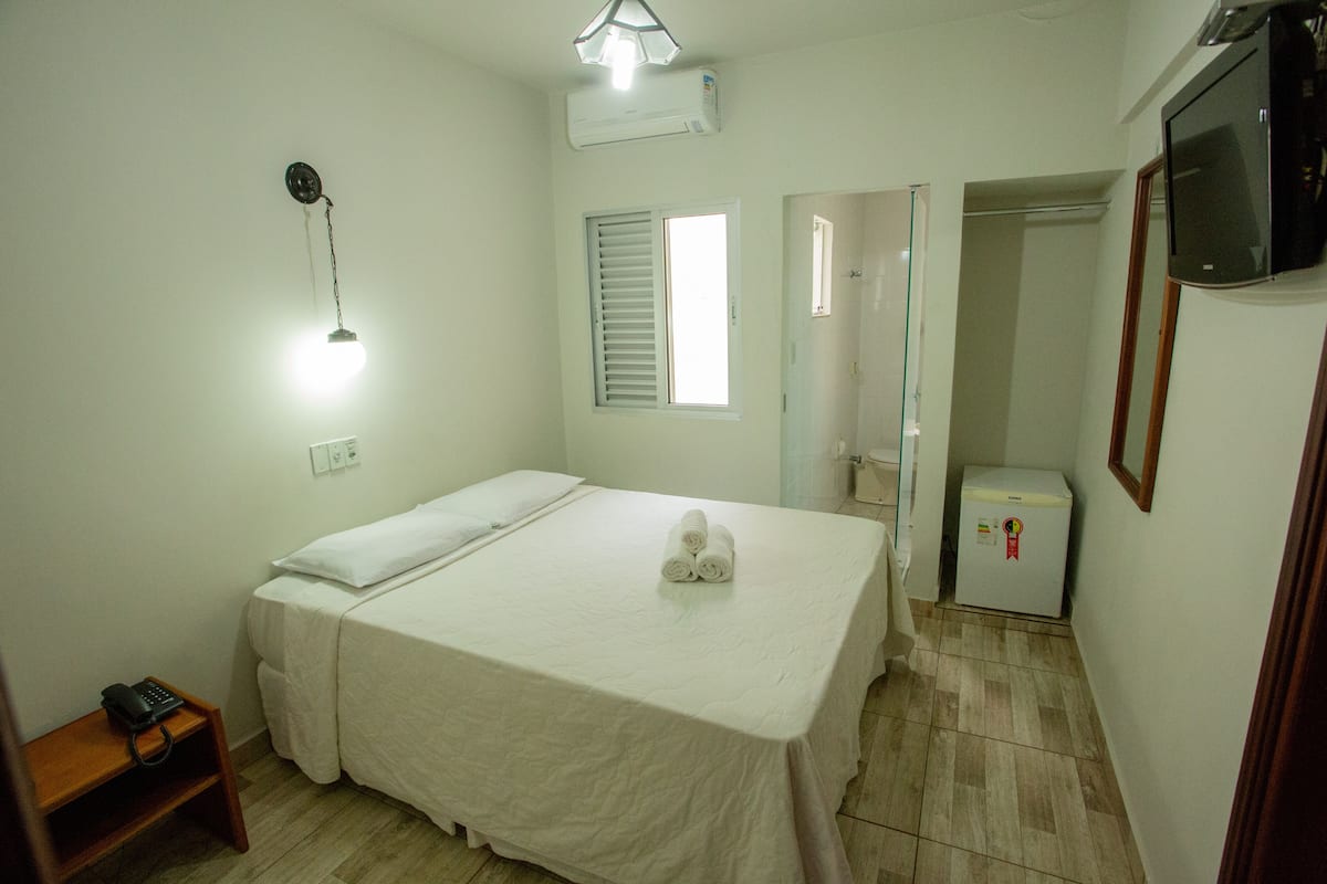 Habitación de hotel con cama, lámpara de noche, mesita, refrigerador y televisor.